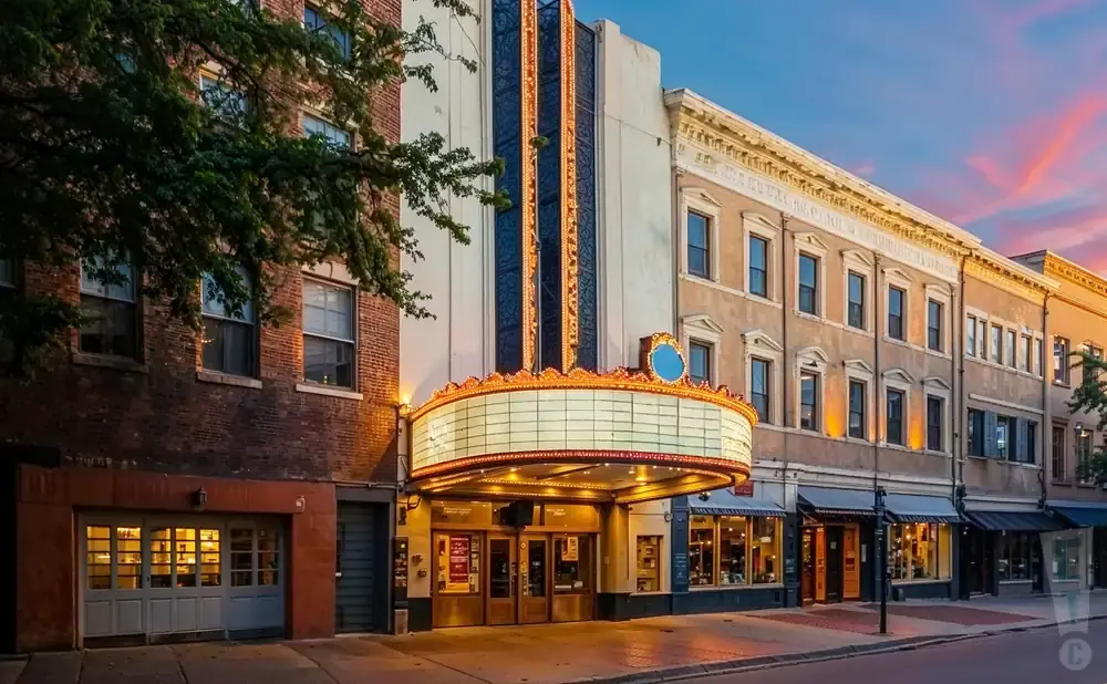 A vintage theater marquee illuminated at dusk on a city street, with brick buildings and shops lining the sidewalk.