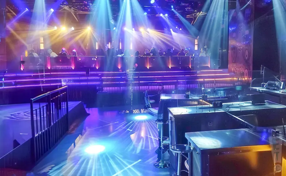 Stage with bright blue and yellow lights, people seated at a long table, and tables with stools in the foreground.