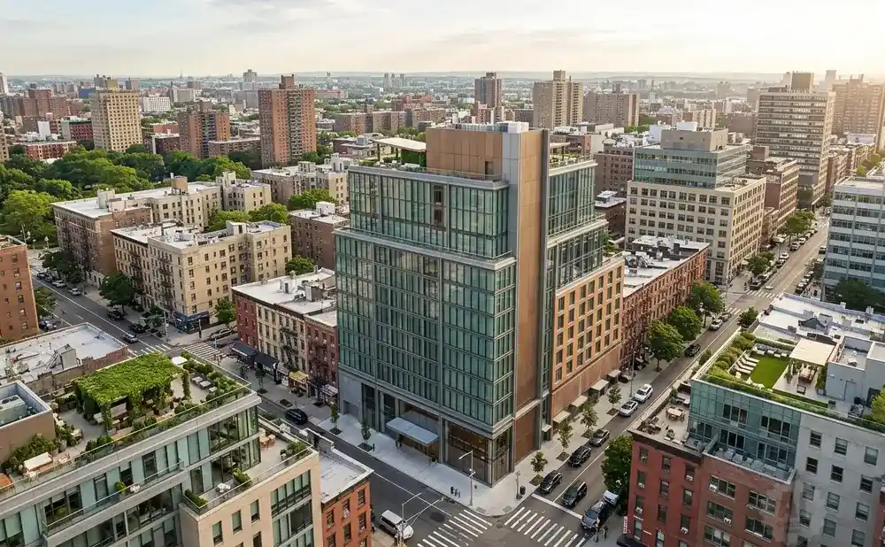 Aerial view of a modern glass building with a green rooftop terrace, surrounded by older brick buildings in a city.