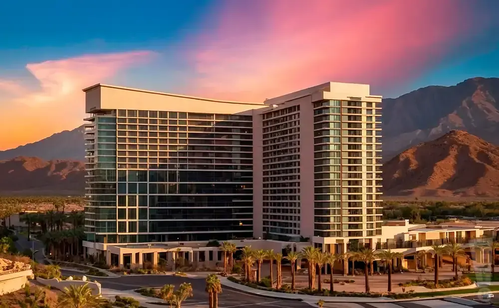 A modern hotel with glass windows stands against a colorful sunset and mountains.