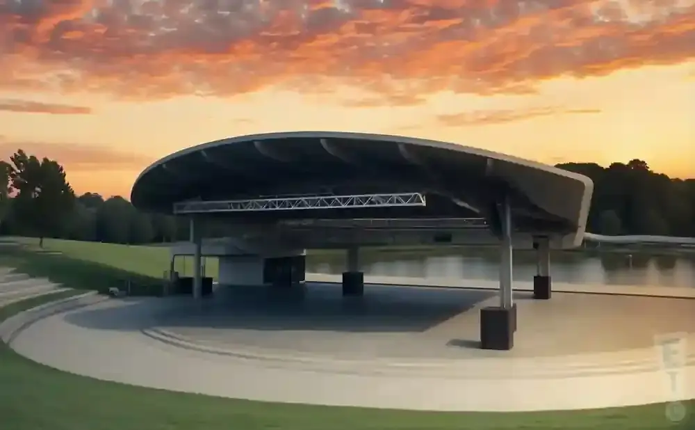 Outdoor amphitheater with a curved roof on a stage, overlooking a lake at sunset.