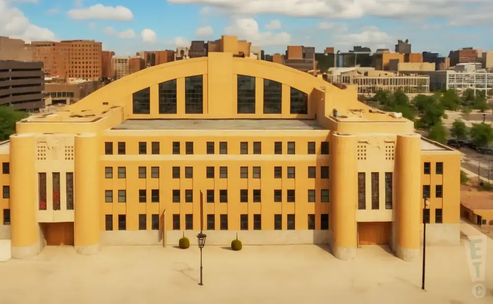 An aerial view of a large, tan building with a distinctive arched roof and numerous windows, set against a backdrop of a city skyline under a cloudy sky.