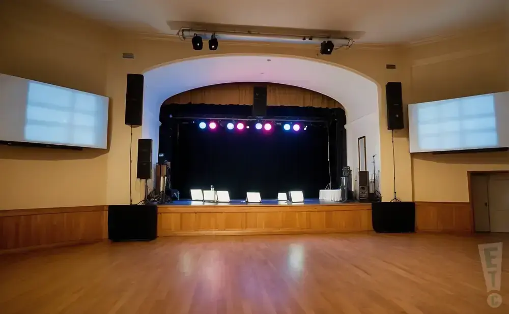 A stage with black curtains, colorful lights, and speakers in an empty wooden-floored room.