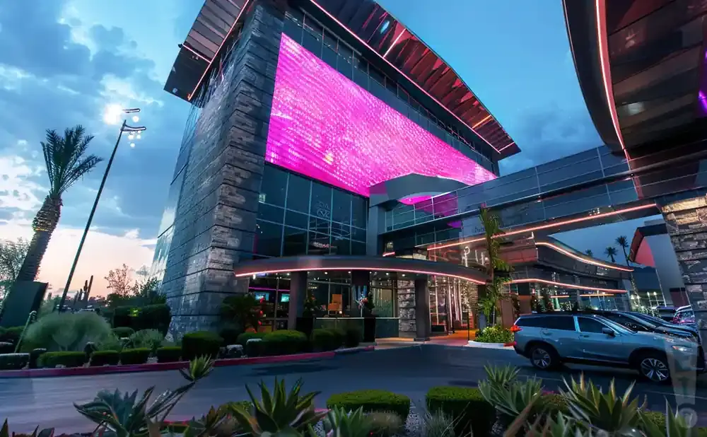 Modern building with a large pink LED screen, palm trees, and cars parked outside at dusk.