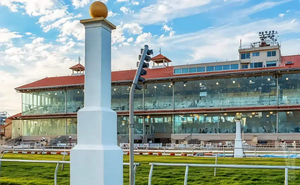 A white column topped with a gold ball stands in front of a modern grandstand at a racetrack under a cloudy sky.