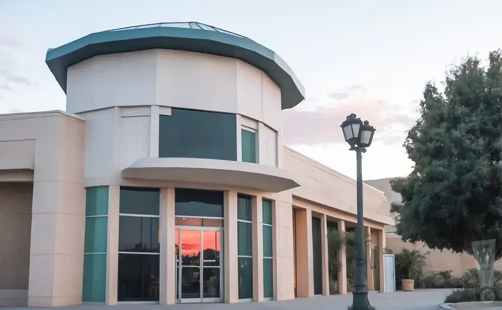 Modern building entrance with glass doors and windows reflecting a sunset, a lamppost, and a large tree.