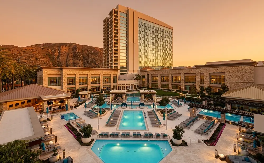 Aerial view of a resort complex with multiple pools, lounge chairs, and a high-rise building at sunset.