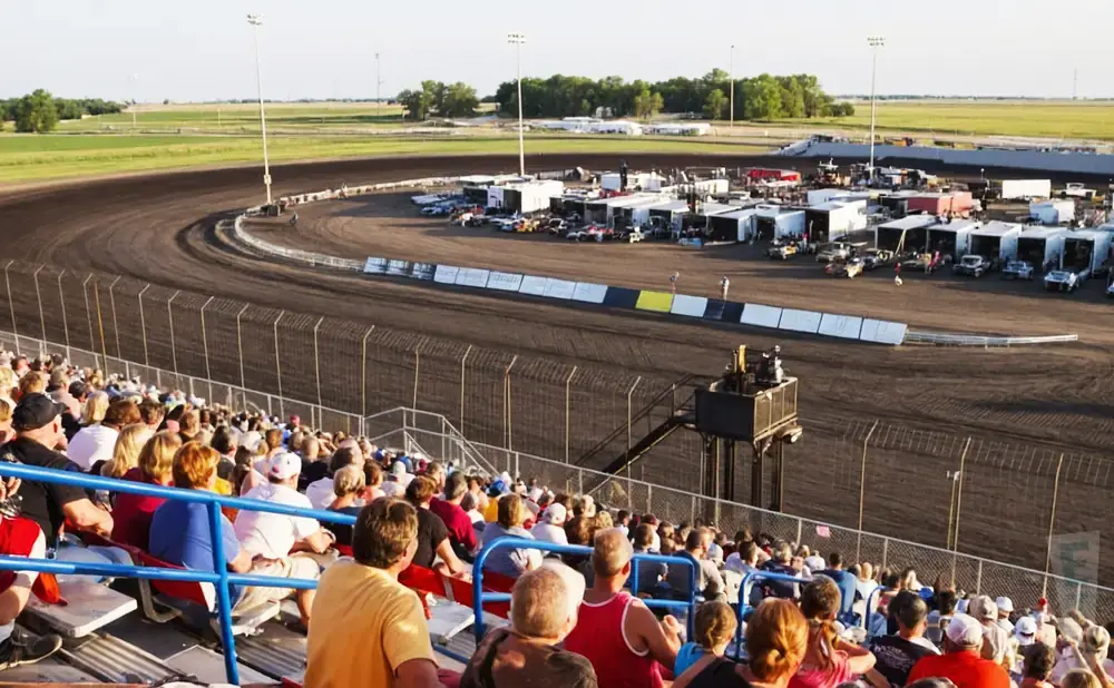 Fans watch a dirt track race at a stadium, with cars lined up in the pit area.