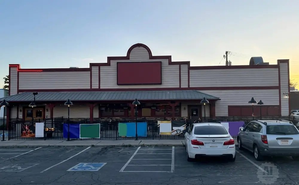 Exterior of a restaurant with a large red sign, patio seating, and cars in the parking lot.