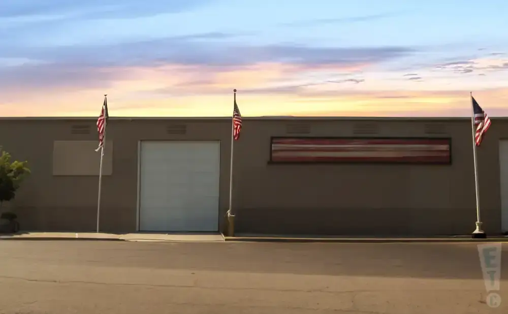 An American flag stands outside a warehouse with a large rollup door and a mural under a sunset sky.