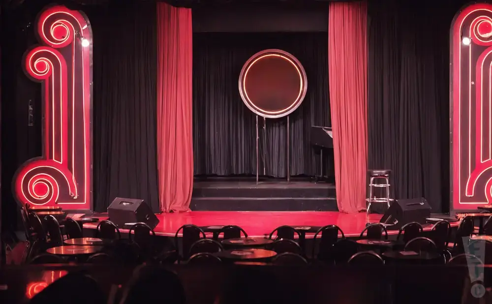 A dimly lit comedy club stage with red curtains, a circular neon sign, and tables in the foreground.