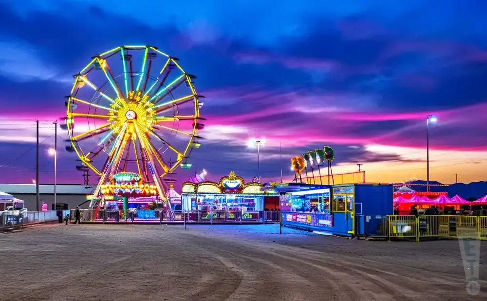 A Ferris wheel and carnival rides are lit up at dusk with a colorful sunset and purple clouds.