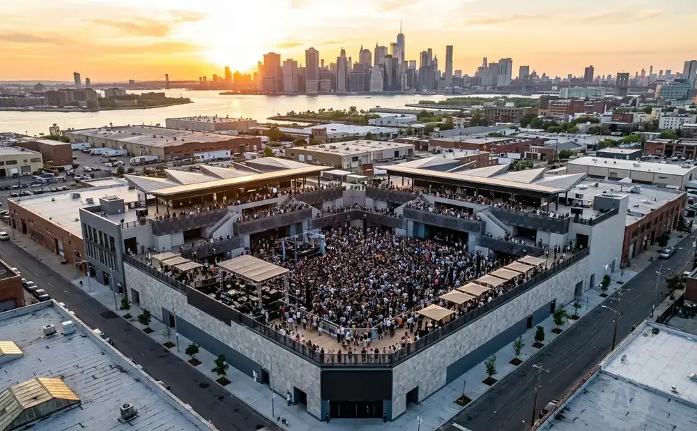 Aerial view of a crowded outdoor concert venue with a city skyline and sunset in the background.