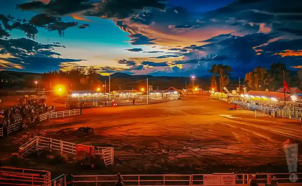Rodeo arena at dusk with dramatic clouds, illuminated grandstands, and bleachers filled with spectators.