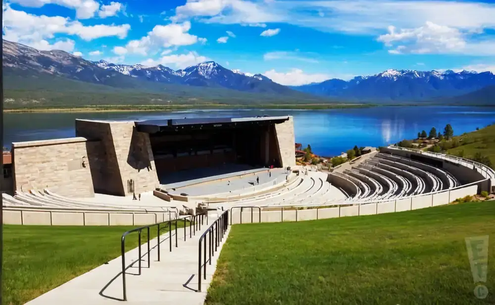 Outdoor amphitheater with stage overlooking a lake and snow-capped mountains under a blue, cloudy sky.