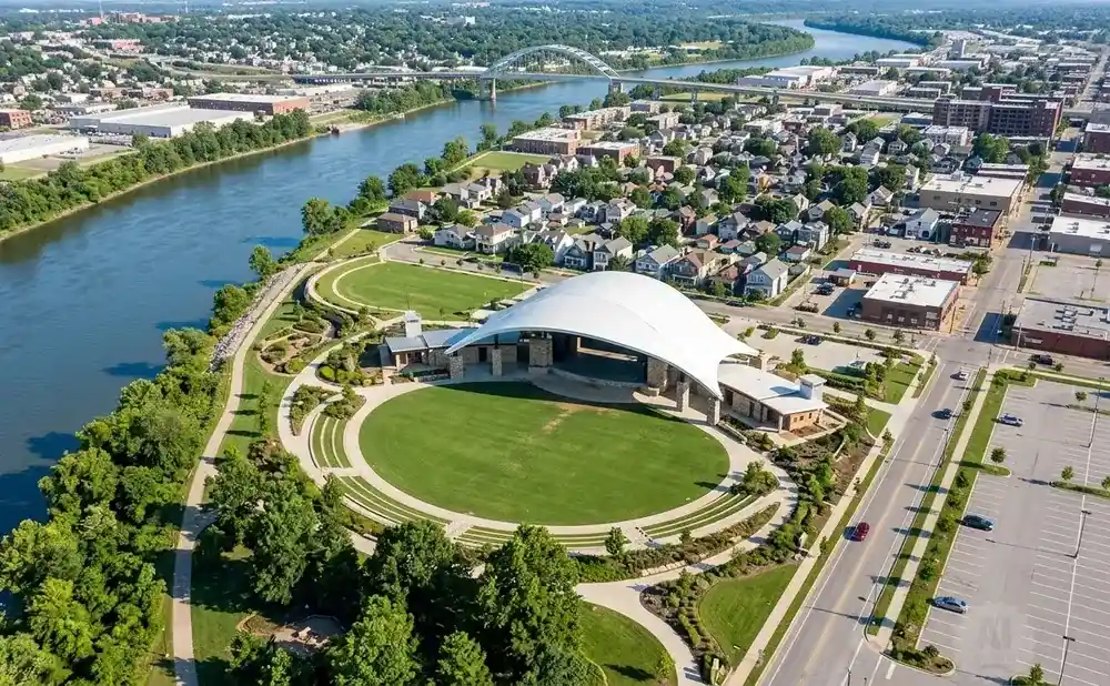 Aerial view of an outdoor amphitheater with a white tent, green lawn, and tiered seating, next to a wide river.
