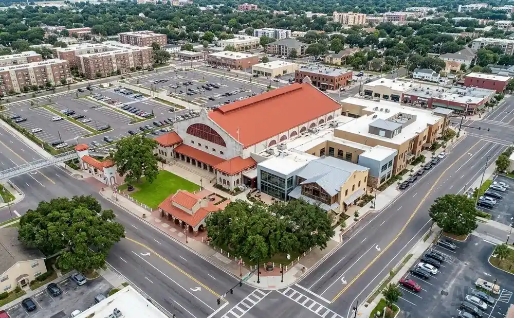Aerial view of a downtown area with large buildings, a red-roofed structure, a parking lot, and trees.