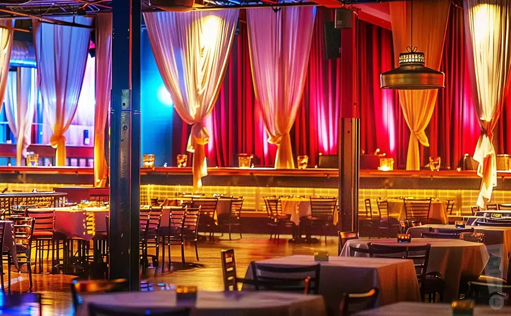 A dimly lit event space with tables set for dining, draped curtains, and stage lighting.