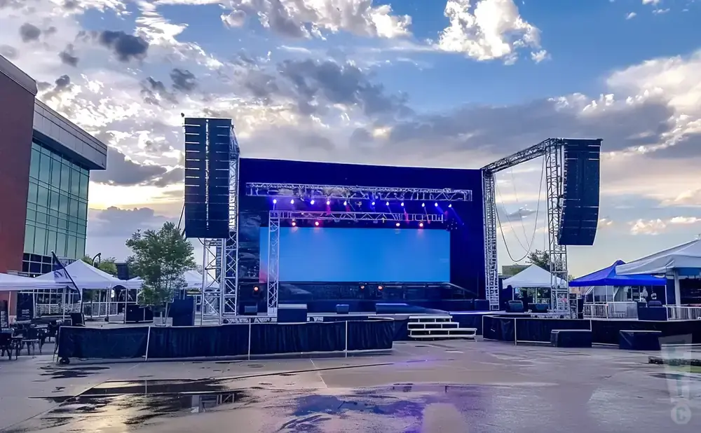 Outdoor stage with large screen, speakers, tents, and a cloudy sky.