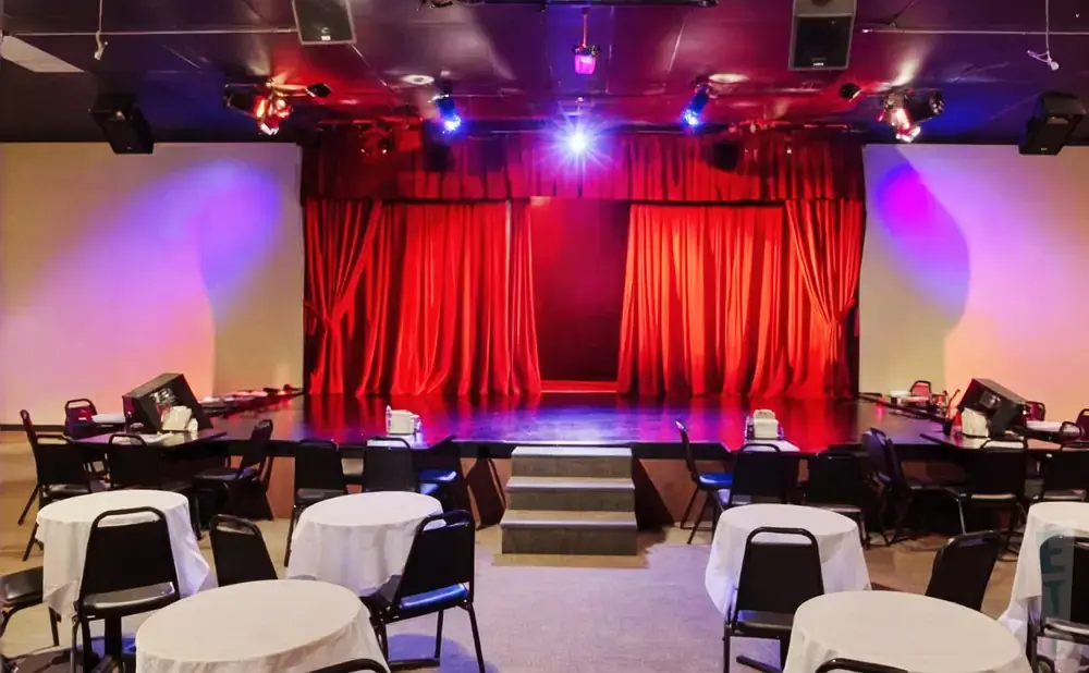 A theater stage with red curtains, spotlight, and tiered seating with tables covered in white cloths.