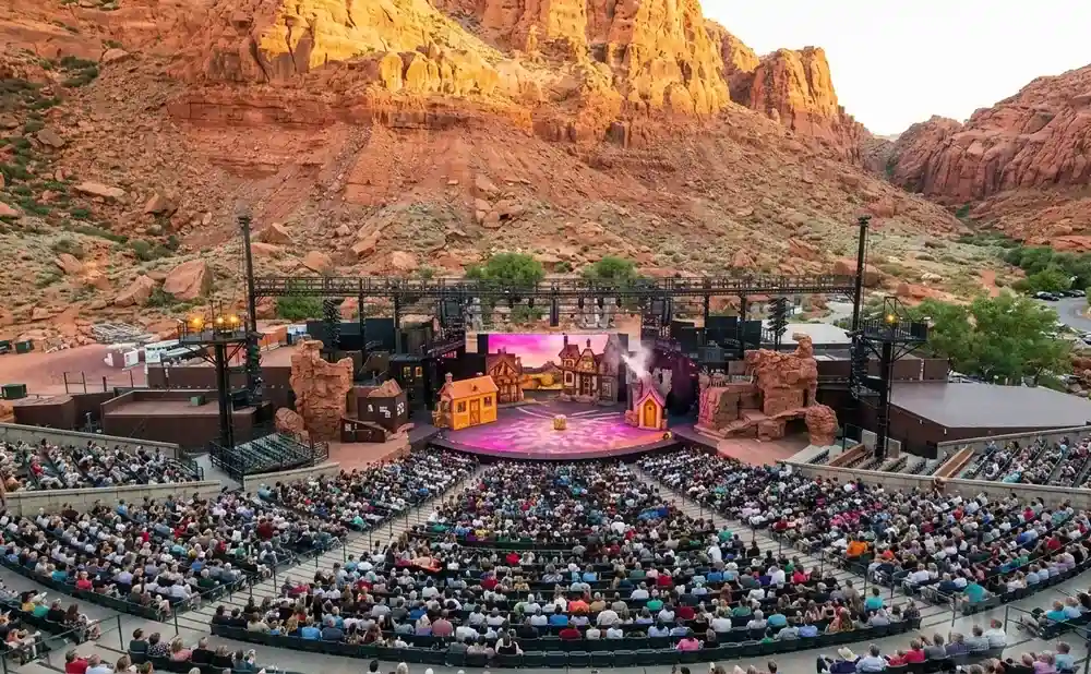 Outdoor theater in a canyon with a stage set with houses and a large audience seated in tiered rows.