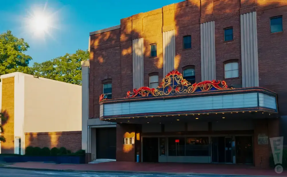 Sunlit brick theater with marquee, flanked by a light-colored building and trees.