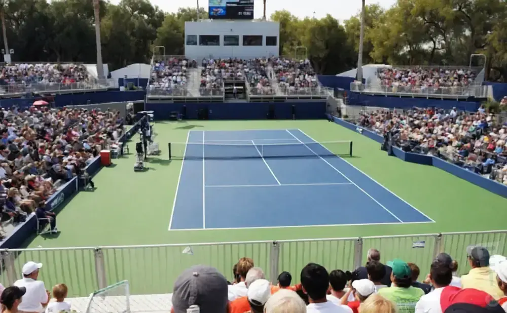 A tennis court is surrounded by bleachers full of spectators under a sunny sky.