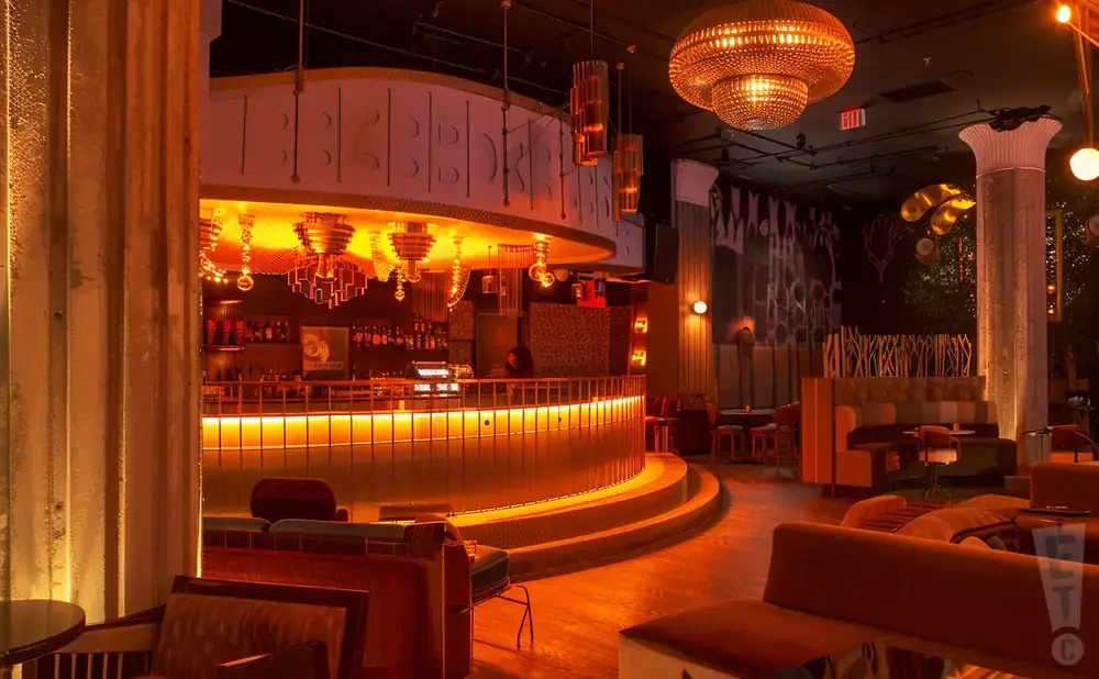 Interior of a dimly lit bar with orange lighting, a curved bar, plush seating, and ornate chandeliers.