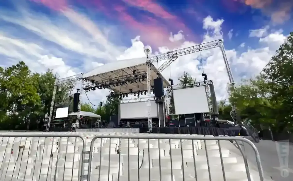 An outdoor concert stage with empty white seats in the foreground under a colorful sky.