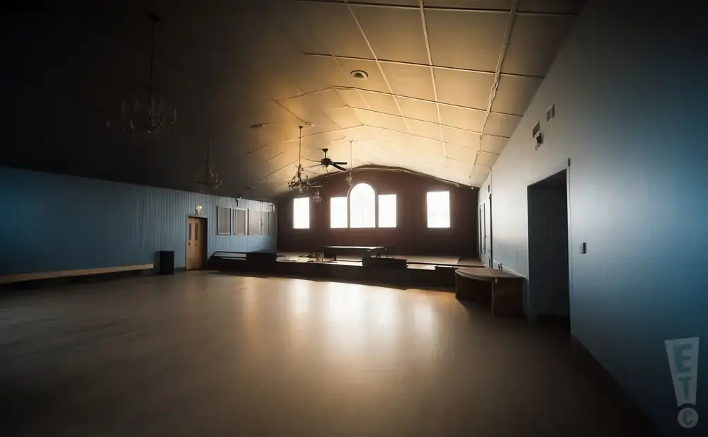 An empty, dimly lit room with a stage, large windows, and a ceiling fan.