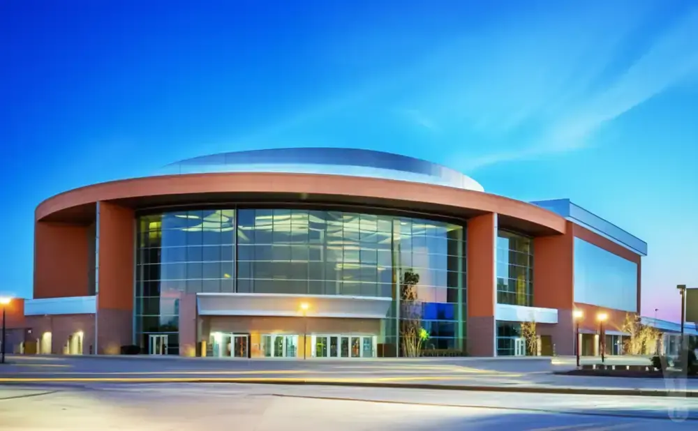 A modern, rounded building with large glass windows and orange exterior walls, illuminated by streetlights under a blue sky.