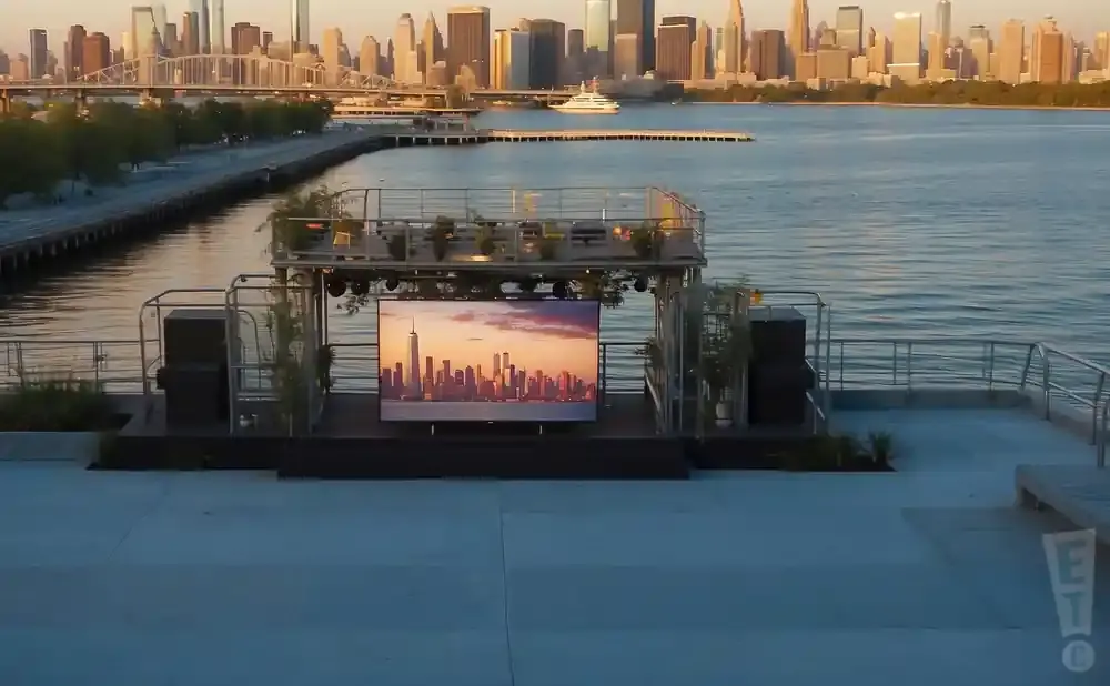 Outdoor stage with a large screen showing a city skyline at sunset, overlooking a body of water.
