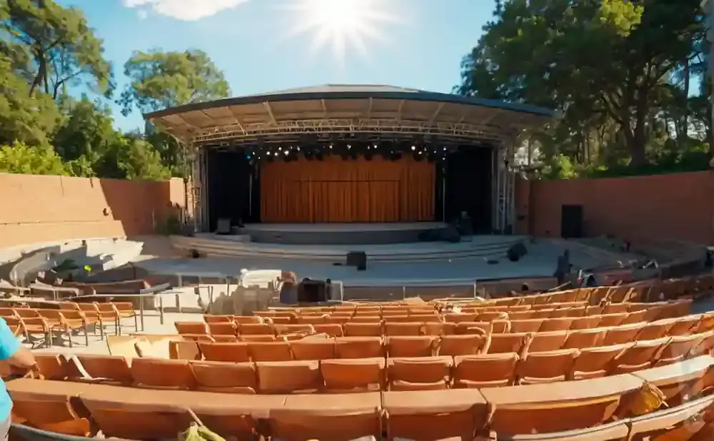 Outdoor amphitheater with rows of empty orange seats facing a stage with a large orange curtain and lights.