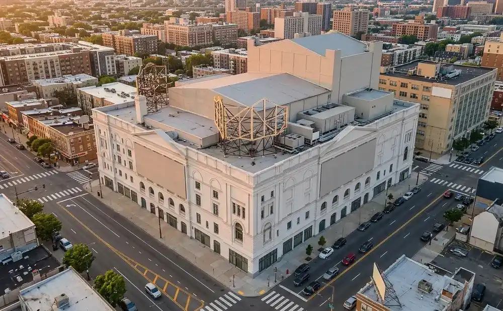 Aerial view of a large white building with a distinctive metal structure on its roof, surrounded by city streets and buildings.