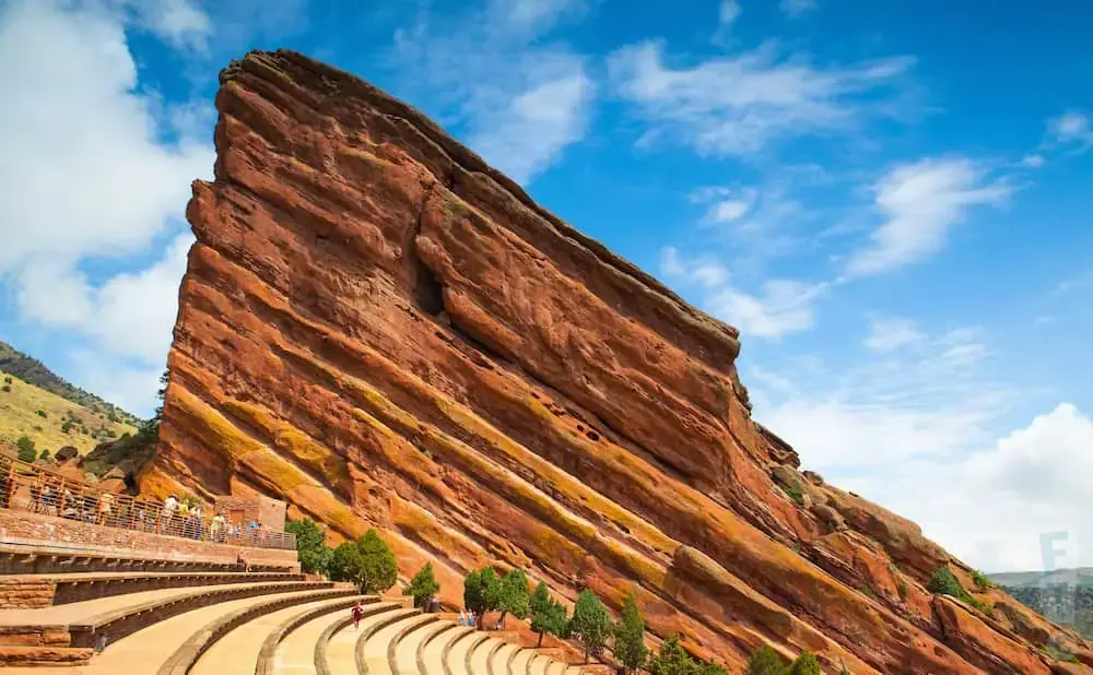 Red Rocks Amphitheatre in Colorado with tiered seating against a massive red rock formation under a blue, cloudy sky.