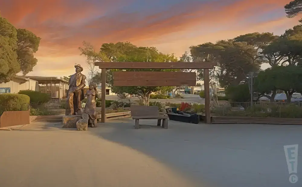 Bronze statues of a man and woman stand near a wooden bench and sign under a colorful sunset sky.