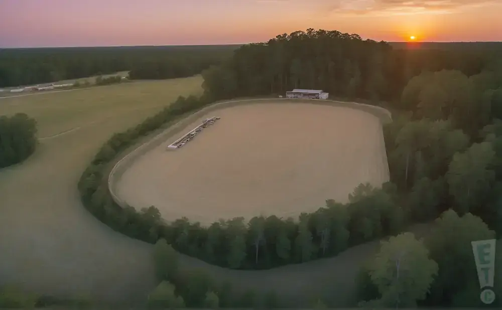 An aerial drone view of a dirt racetrack surrounded by trees at sunset.