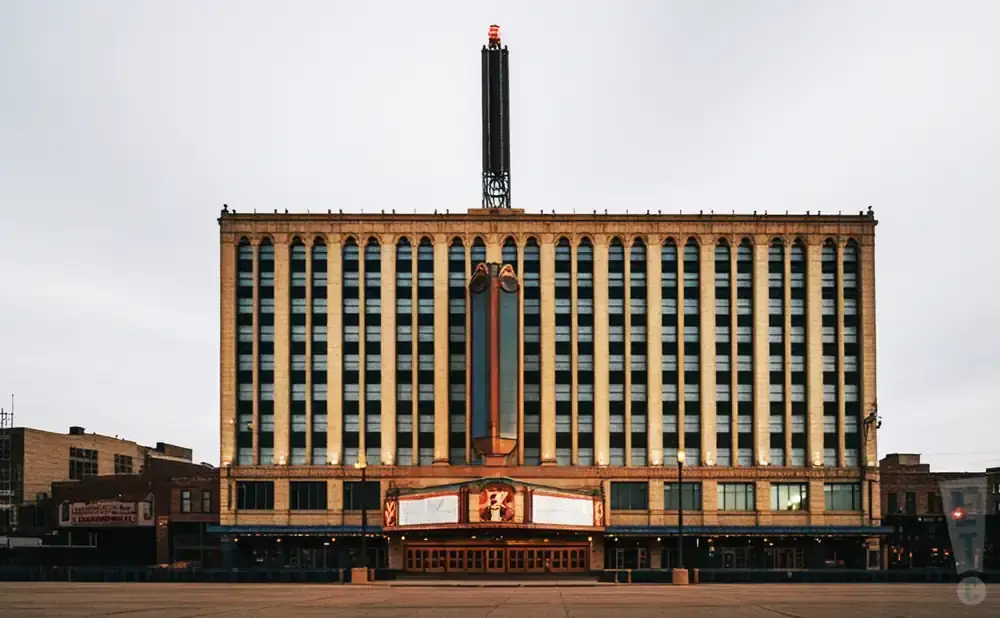 Grand theater building with an illuminated marquee and tall spire against a cloudy sky.