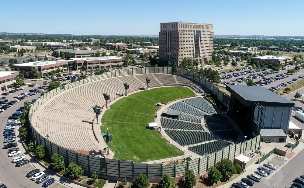 Aerial view of an outdoor amphitheater with tiered seating, a grassy field, and a stage, surrounded by parking lots and office buildings.