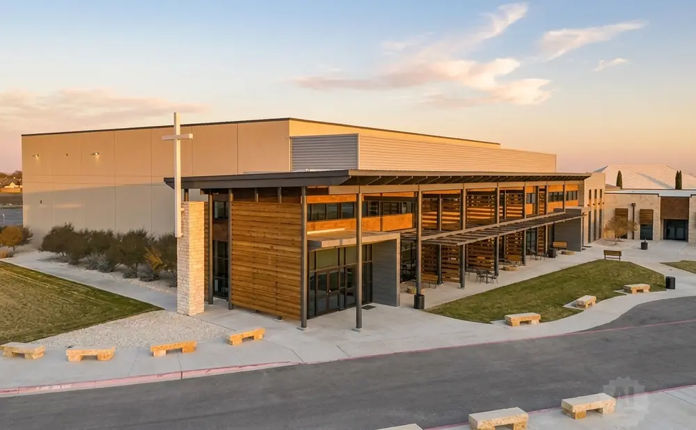 Modern church building with a large white cross, wooden accents, and outdoor seating areas bathed in warm sunset light.