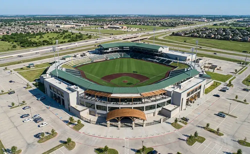 Aerial view of a baseball stadium with surrounding parking lots and residential areas under a clear blue sky.
