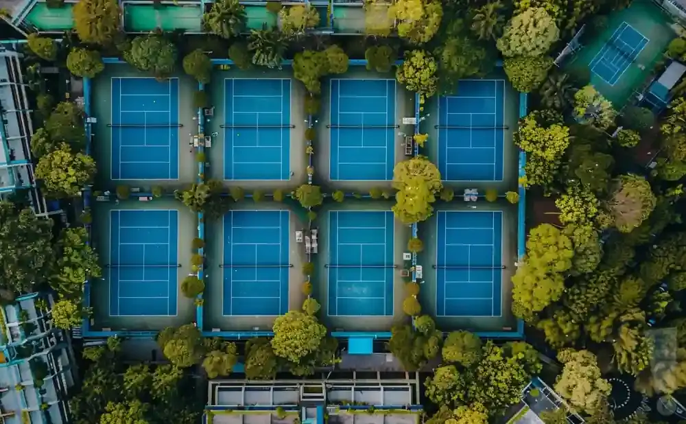 Aerial view of multiple blue tennis courts surrounded by lush green trees and foliage.