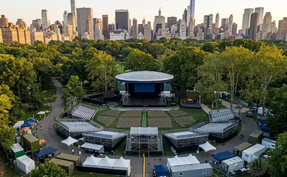 Aerial view of an outdoor amphitheater with tiered seating, a stage, and a city skyline in the background.