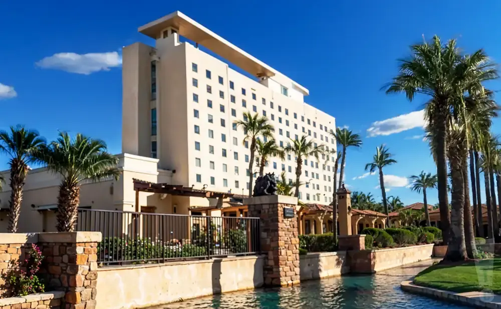 A tall hotel with many windows stands behind palm trees, with a water channel in the foreground.