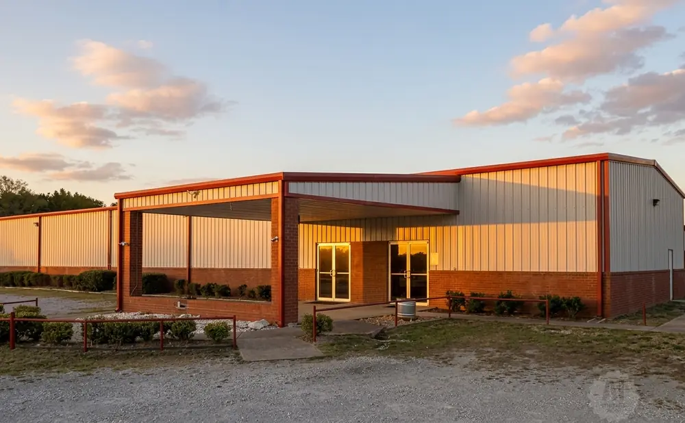 A modern building with white corrugated metal siding and red trim, lit by warm sunset light.