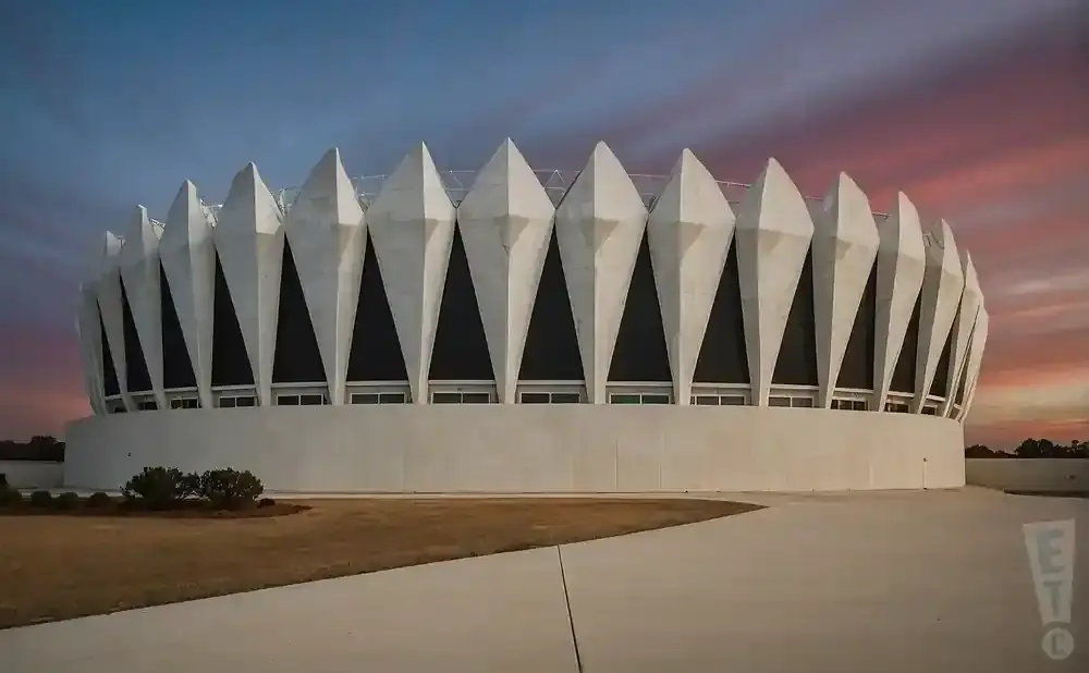 A modern, circular building with pointed white accents and dark windows against a sunset sky.