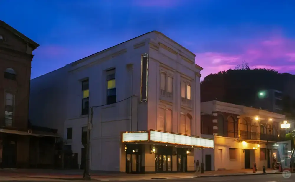 A historic theater with a lit marquee stands on a street at twilight, with a purple and blue sky above.