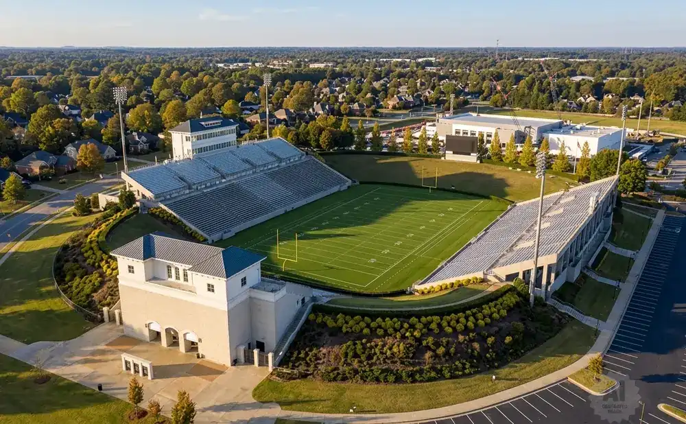 Aerial view of a football stadium with green field, bleachers, and surrounding buildings under a blue sky.