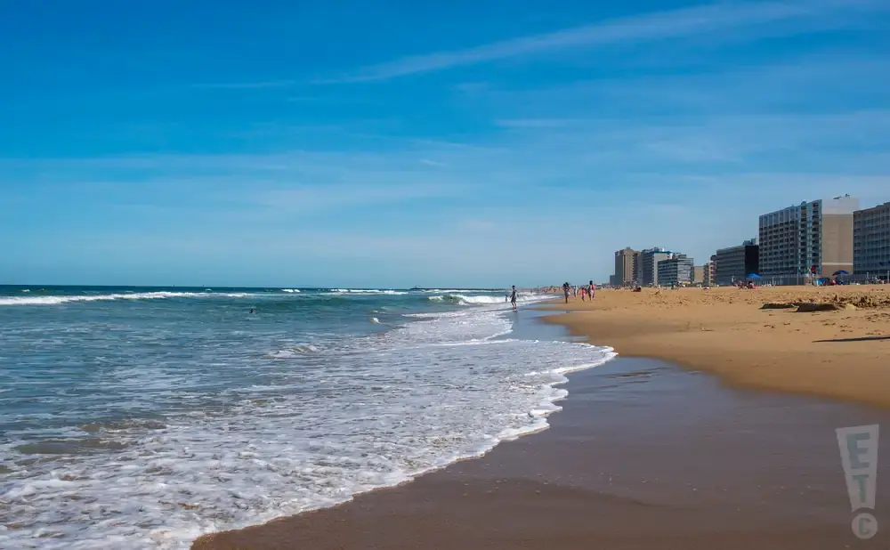 A wide beach with ocean waves lapping the shore, backed by hotels under a bright blue sky.