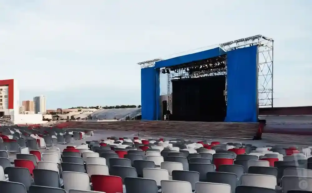 An outdoor amphitheater with a stage, seating, and a city skyline in the background.
